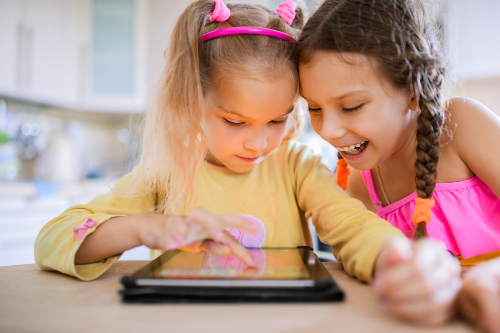 Na imagem, duas meninas interagem com um tablet. A imagem é usada para ilustrar a importância do conteúdo digital interativo no ensino e na aprendizagem.