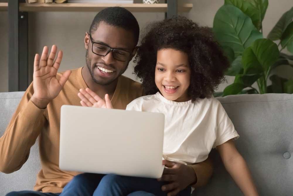 Na imagem, um pai e uma filha criança, assistem uma aula online juntos. Ambos interagem levantando as mãos. A imagem foi usada para ilustrar o artigo sobre as particularidades do conteúdo digital para anos iniciais.