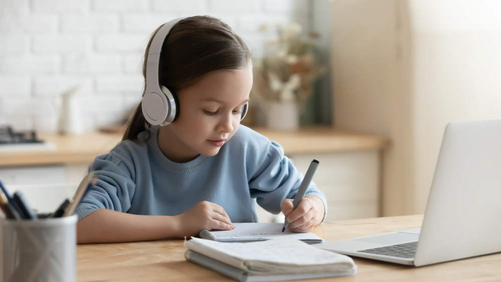 A imagem mostra uma criança estudando de casa, escrevendo em um caderno enquanto escuta algo com fones de ouvido. A imagem ilustra a temática do artigo sobre o ensino híbrido nos anos iniciais.