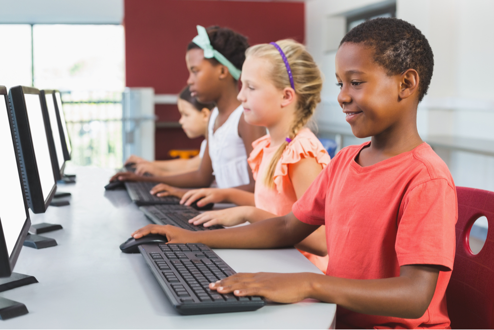 A imagem traz crianças em uma sala, mexendo em computadores. A imagem tem como objetivo ilustrar a aplicação de objetos interativos nos anos iniciais de ensino.