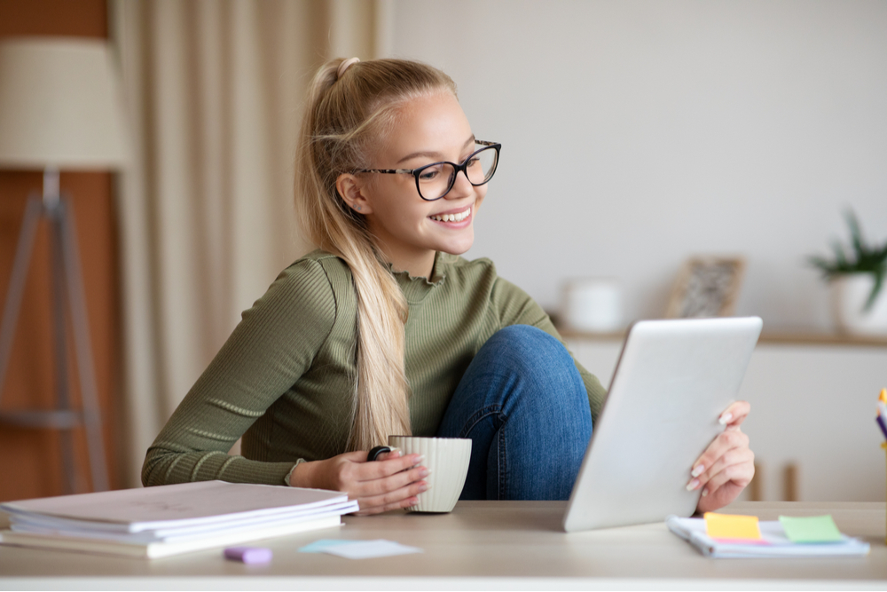A imagem traz uma adolescente, em casa, assistindo algo em um tablet. Ela está sorrindo e segurando uma xícara. A imagem foi usada para ilustrar o artigo sobre como utilizar o digital no Novo Ensino Médio.