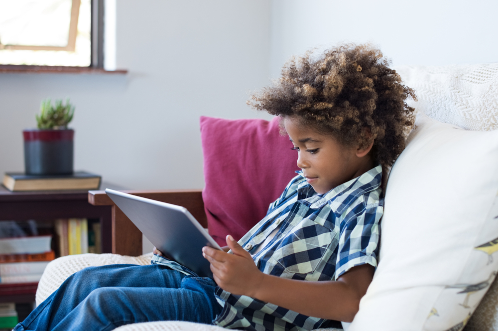 Na imagem, uma criança mexe em um tablet sentada no sofá. O objetivo da imagem é ilustrar o artigo sobre os melhores formatos de objetos interativos para Anos Iniciais.